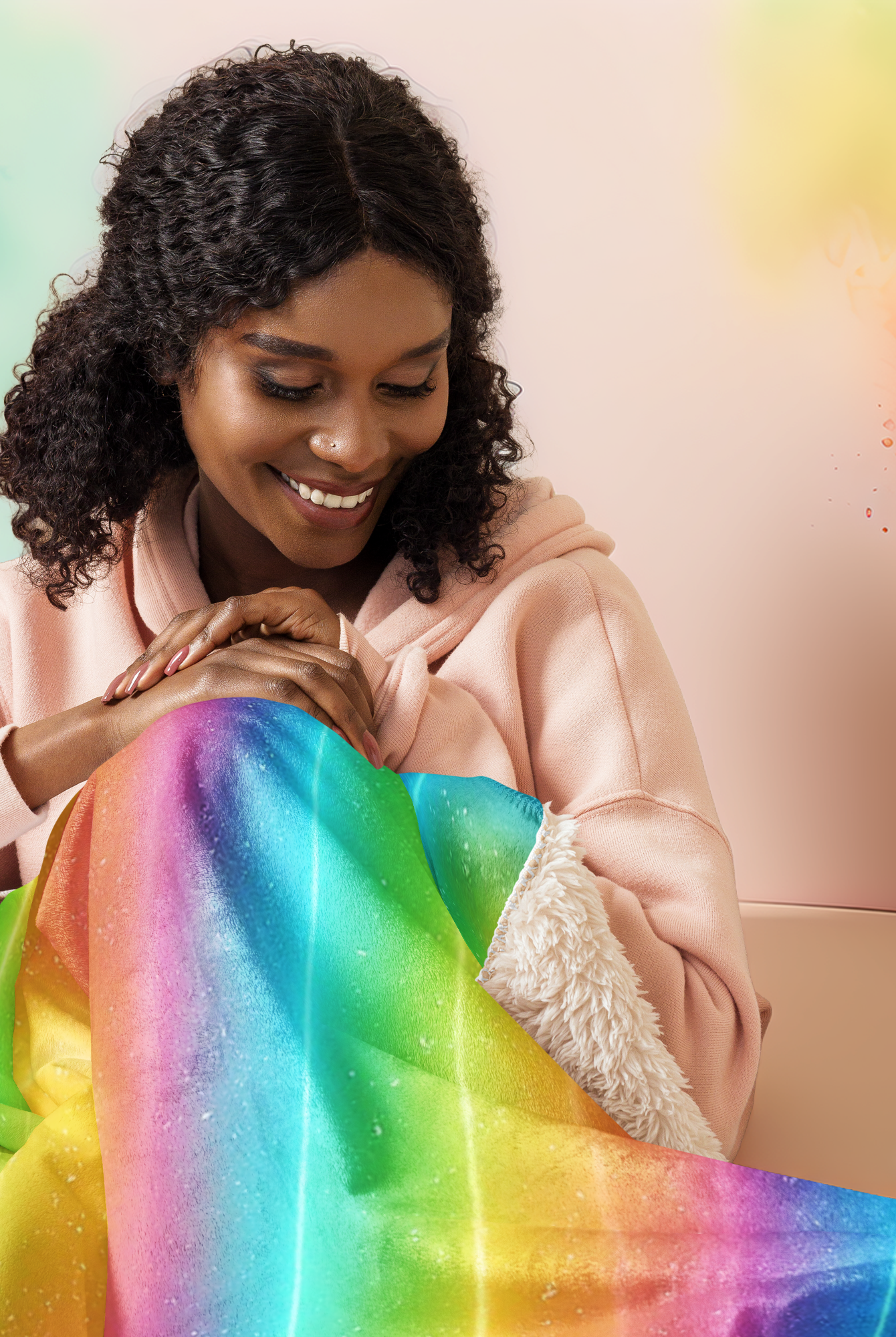 Femme assise et souriante, enveloppée dans une couverture sherpa arc-en-ciel brillante, dans un décor lumineux aux tons pastel.