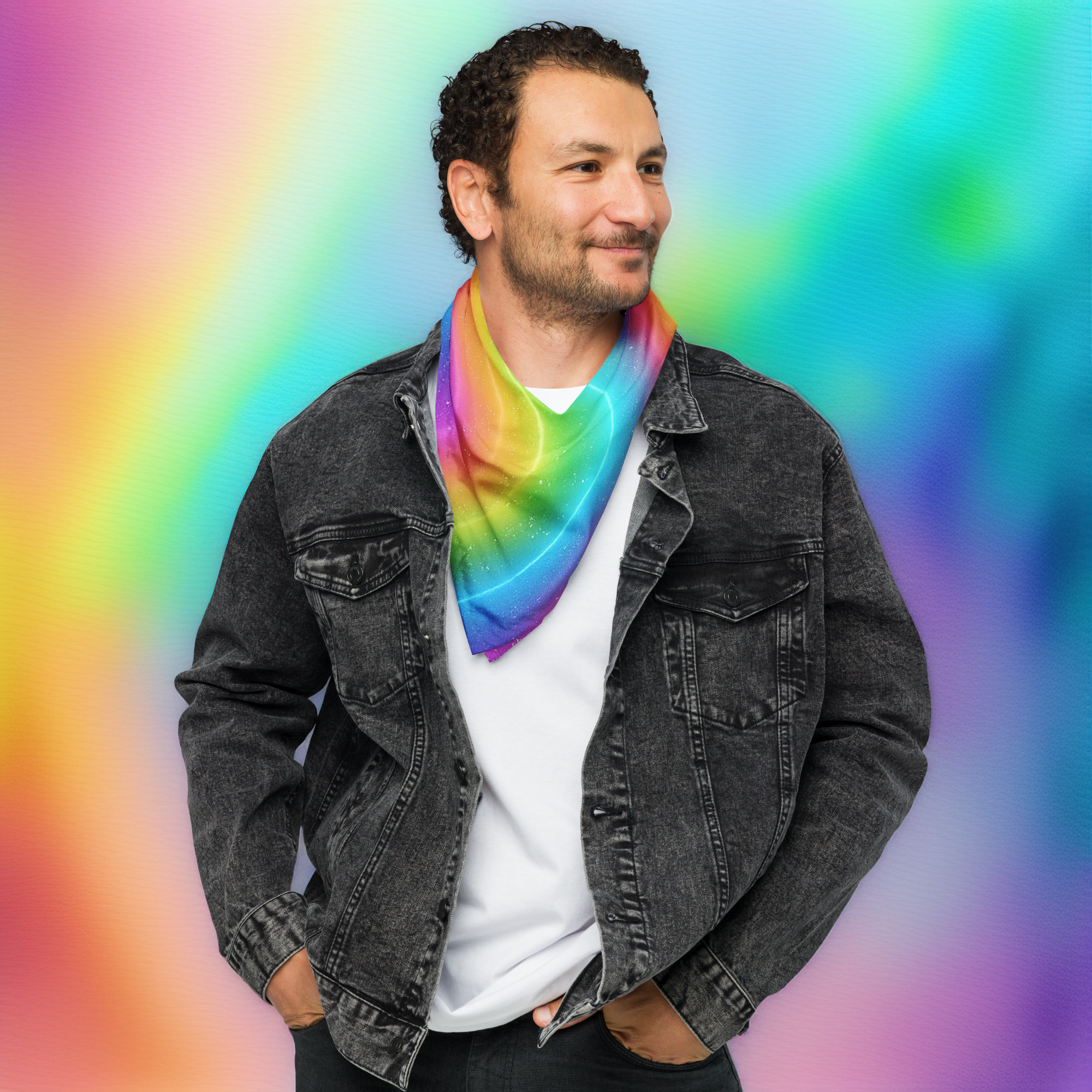 Portrait d’un homme souriant portant un bandana arc-en-ciel noué autour du cou, debout en veste en jean noire devant un fond coloré dégradé aux tons pastel.